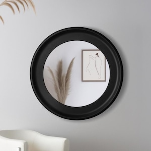 Może przedstawiać: Okrągłe czarne lustro wisi na szarej ścianie, odbijając oprawione dzieło sztuki i suszone rośliny. Białe krzesło z chromowaną ramą znajduje się poniżej, tworząc minimalistyczną estetykę. Rama lustra jest gruba i matowa.