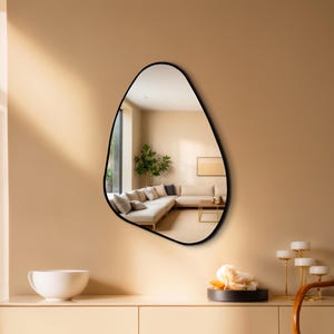 May include: An irregular, organic-shaped wall mirror featuring a thin, smooth black frame. The reflective surface displays a clear view of a living room interior, including a beige sectional sofa, a wooden coffee table, and a green potted plant.