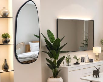 Puede incluir: Una cómoda blanca con tres cajones y un espejo enmarcado en negro encima. Una gran planta en maceta con hojas verdes está delante de la cómoda. Un espejo ovalado negro con un borde ondulado está en la pared a la izquierda de la cómoda.