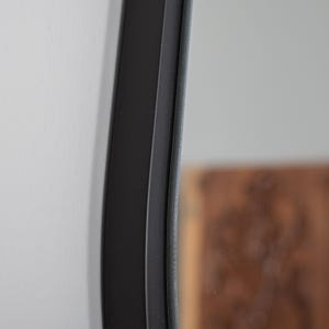 May include: A close-up view of a circular mirror featuring a smooth, matte black frame. The frame has a distinct, slightly curved profile that extends outward from the reflective glass surface, creating a clean and defined edge against the wall.