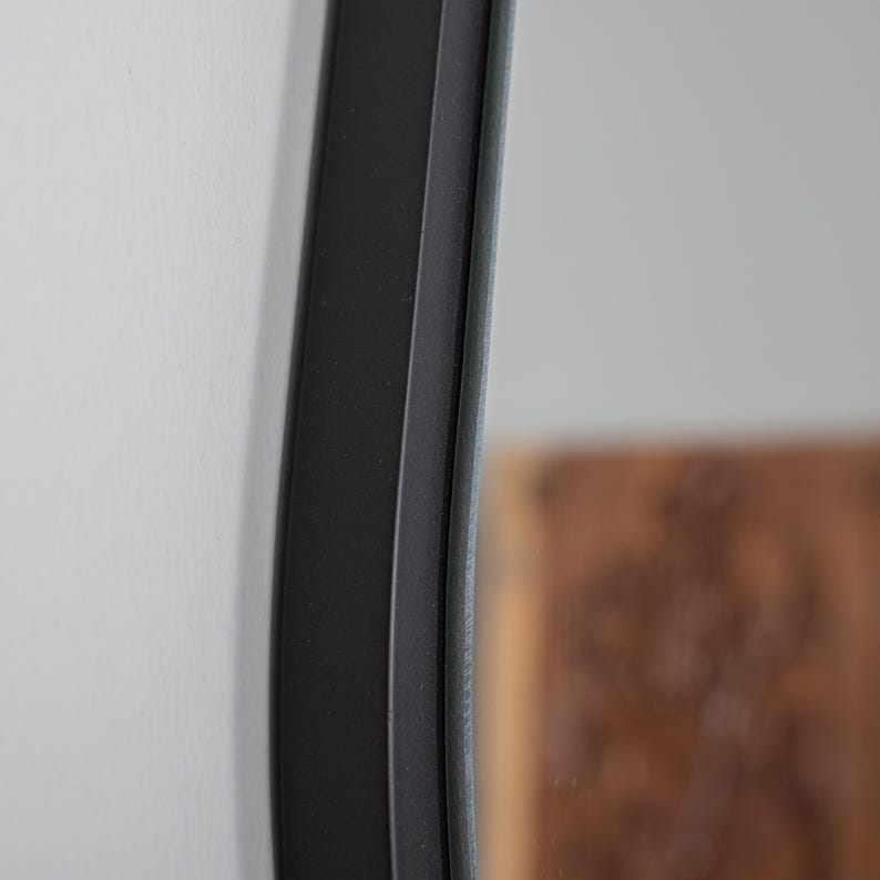 May include: A close-up view of a circular mirror featuring a smooth, matte black frame. The frame has a distinct, slightly curved profile that extends outward from the reflective glass surface, creating a clean and defined edge against the wall.