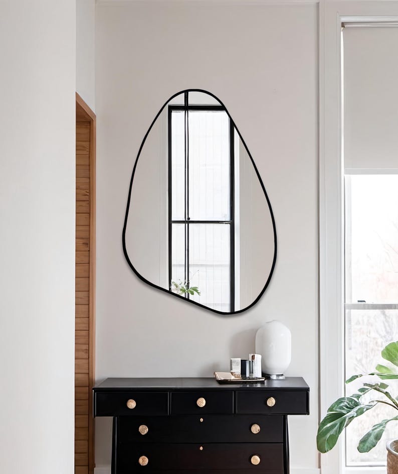 Modern black asymmetrical wall mirror with thin edge, mounted above console in Japandi-style entryway.