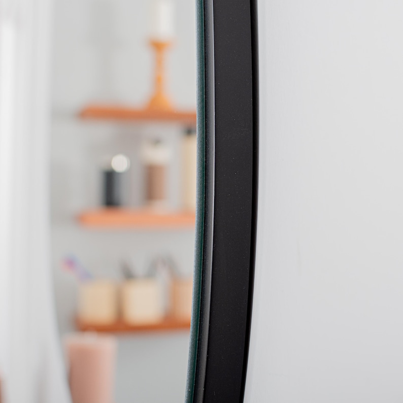 May include: A circular mirror featuring a smooth, matte black frame. The reflective glass surface is positioned vertically, capturing a blurred reflection of wooden shelves and various household items in the background, emphasizing the frame's edge.