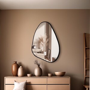 May include: An asymmetrical wall mirror with a thin black frame featuring an irregular, organic shape. The reflective surface displays a room interior with a tall plant in a pot, capturing a clear view of the surroundings within its curved border.
