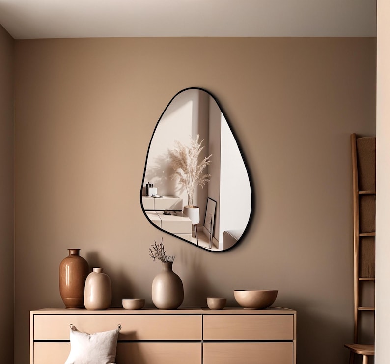 May include: An asymmetrical wall mirror with a thin black frame featuring an irregular, organic shape. The reflective surface displays a room interior with a tall plant in a pot, capturing a clear view of the surroundings within its curved border.