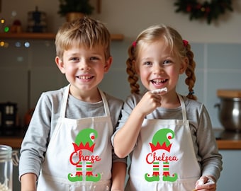 Puede incluir: Dos niños con delantales blancos con diseños de elfos y nombres en rojo. Los delantales tienen sombreros y pies de elfo verdes. Los niños sonríen y parecen estar horneando en una cocina.