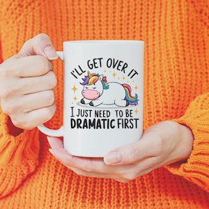 May include: White ceramic mug with the text "I'll get over it, I just need to be dramatic first" and a cartoon unicorn with a rainbow mane and tail. The mug is held by a person wearing an orange sweater.