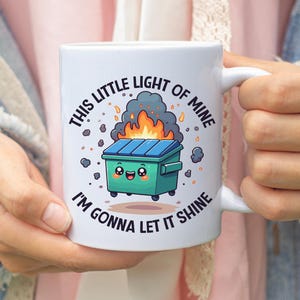 May include: White ceramic mug featuring a cartoon dumpster on fire with the text "THIS LITTLE LIGHT OF MINE I'M GONNA LET IT SHINE" in a circular design. The dumpster has a smiling face.
