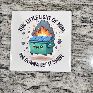 May include: Square white tile with a cartoon illustration of a teal dumpster on wheels with a smiling face, flames, and the text "THIS LITTLE LIGHT OF MINE I'M GONNA LET IT SHINE" in black.