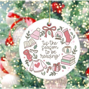 May include: Round white Christmas ornament with the text "tis the season to be reading" surrounded by illustrations of books, a sweater, a hat, a scarf, and other winter-themed items. The ornament hangs from a gold string.
