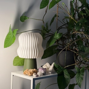 Può includere: Una lampada in ceramica bianca a coste con base verde è appoggiata su una mensola in metallo bianco. La lampada è accanto a una varietà di cristalli e a una pianta in vaso con grandi foglie verdi. Il design della lampada è moderno e scultoreo.
