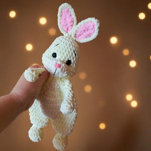 May include: A cream-colored crochet rabbit plush toy with long ears featuring pink inner sections. The stuffed animal has black safety eyes, a small pink nose, and a stitched mouth. The texture is soft and chunky, crafted from yarn.