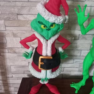 Può includere: Una figura verde del Grinch che indossa un cappello da Babbo Natale rosso con bordino di pelliccia bianca e un abito da Babbo Natale rosso e bianco con una cintura nera. Il Grinch è in piedi con le braccia incrociate e ha un'espressione scontrosa sul viso.