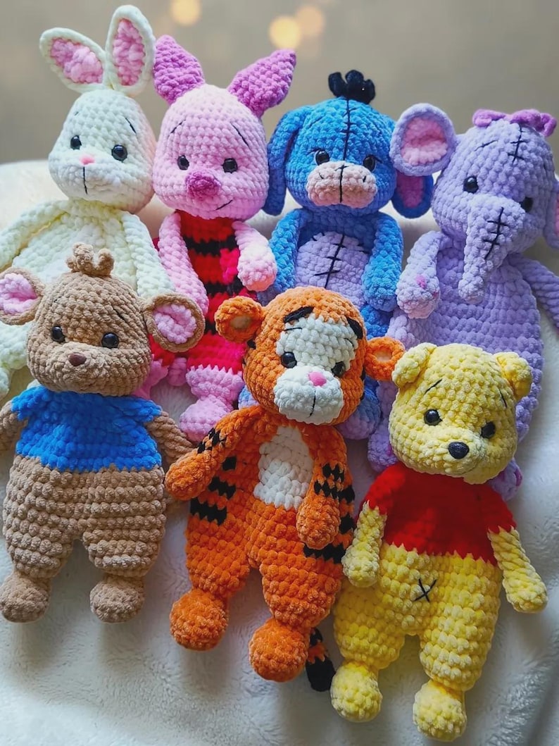 May include: A collection of seven handmade crochet plush toys featuring various animal characters. The group includes a cream rabbit, a pink pig, a blue donkey, a purple elephant, a brown bear, an orange tiger, and a yellow bear, all with black eyes.
