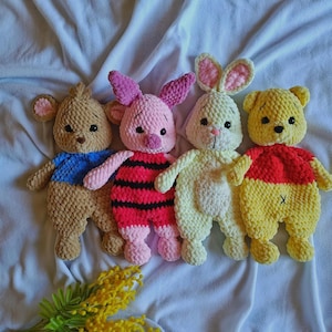 May include: Four plush crochet toys arranged in a row. From left to right: a brown bear with a blue shirt, a pink pig with red and black stripes, a cream-colored rabbit with pink inner ears, and a yellow bear with a red shirt and an X stitch.