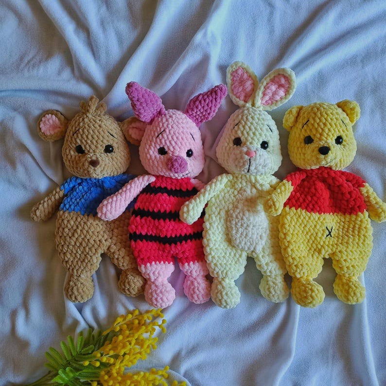 May include: Four plush crochet toys arranged in a row. From left to right: a brown bear with a blue shirt, a pink pig with red and black stripes, a cream-colored rabbit with pink inner ears, and a yellow bear with a red shirt and an X stitch.