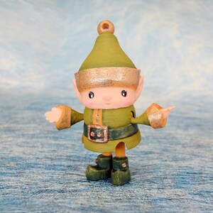 May include: A handcrafted Christmas elf figurine with a green outfit, hat, and boots. The elf has a gold-colored belt buckle and trim on the hat and sleeves. The background is a light blue and white textured surface.