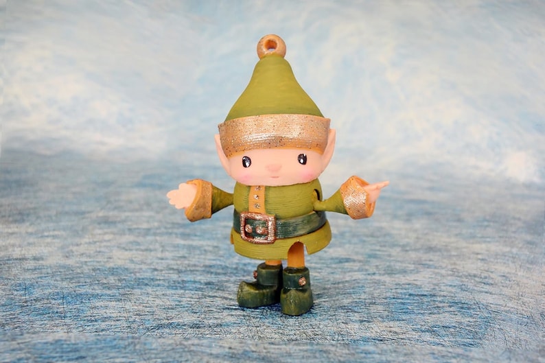 May include: A handcrafted Christmas elf figurine with a green outfit, hat, and boots. The elf has a gold-colored belt buckle and trim on the hat and sleeves. The background is a light blue and white textured surface.