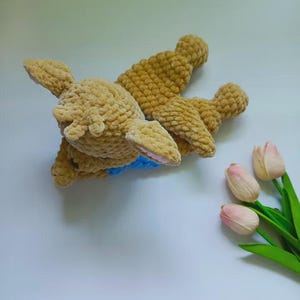 May include: A tan crochet plush toy shaped like a deer with a textured, fuzzy surface, featuring small pink inner ears and a bright blue collar around its neck. The toy is lying down and is positioned next to three light pink artificial tulips.