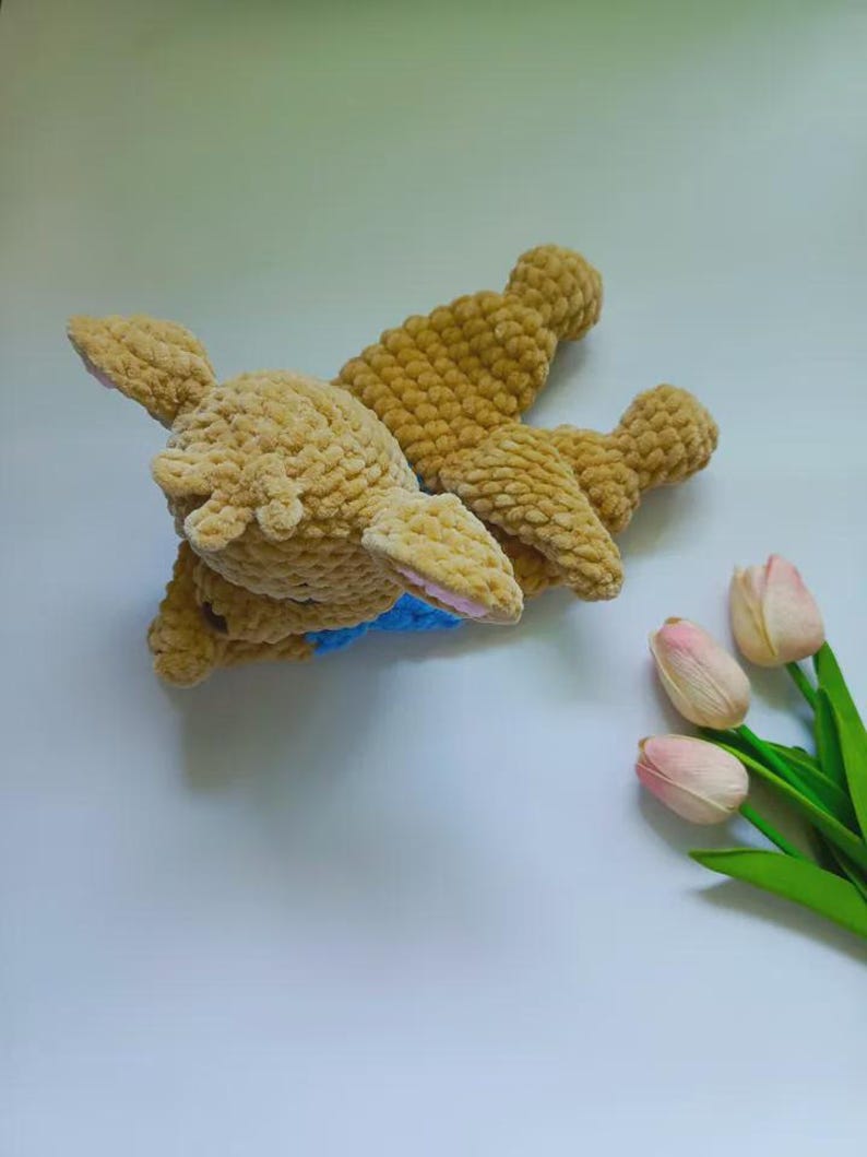 May include: A tan crochet plush toy shaped like a deer with a textured, fuzzy surface, featuring small pink inner ears and a bright blue collar around its neck. The toy is lying down and is positioned next to three light pink artificial tulips.