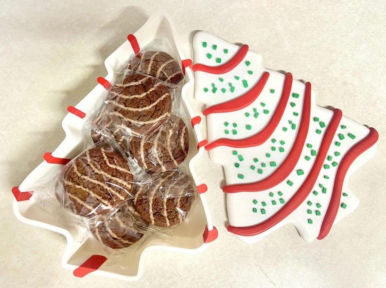 May include: A Christmas tree-shaped serving dish with a lid, filled with individually wrapped chocolate cookies. The lid is decorated with red and green accents. The cookies have a brown and white striped pattern.