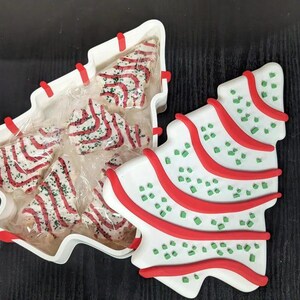 May include: Christmas-themed ceramic serving dish and matching decorative tree. The white dish is shaped like a Christmas tree and contains individually wrapped tree-shaped cookies with red and white stripes. The decorative tree is white with red stripes and green sprinkles.