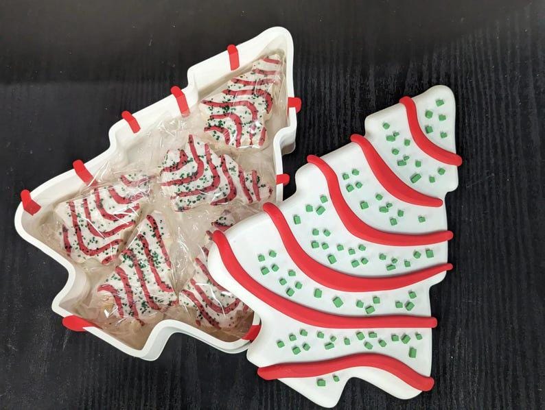 May include: Christmas-themed ceramic serving dish and matching decorative tree. The white dish is shaped like a Christmas tree and contains individually wrapped tree-shaped cookies with red and white stripes. The decorative tree is white with red stripes and green sprinkles.