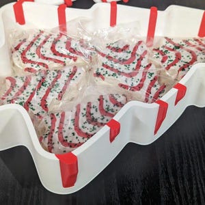 May include: A white, tree-shaped serving tray with red accents holds individually wrapped Christmas tree-shaped cookies. The cookies are decorated with red icing stripes and green sprinkles, creating a festive holiday treat.