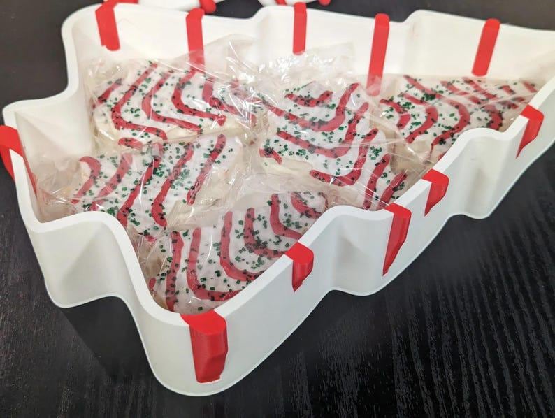 May include: A white, tree-shaped serving tray with red accents holds individually wrapped Christmas tree-shaped cookies. The cookies are decorated with red icing stripes and green sprinkles, creating a festive holiday treat.