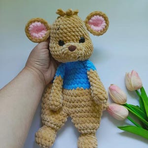 May include: A handmade crochet plush toy shaped like a bear with tan yarn, featuring a bright blue shirt, round ears with pink centers, a dark brown nose, and black safety eyes. The soft, textured yarn creates a thick, looped appearance throughout.