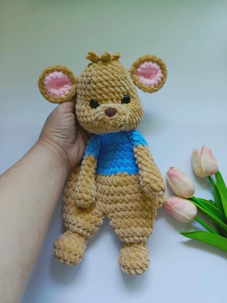 May include: A handmade crochet plush toy shaped like a bear with tan yarn, featuring a bright blue shirt, round ears with pink centers, a dark brown nose, and black safety eyes. The soft, textured yarn creates a thick, looped appearance throughout.