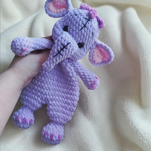 May include: A lavender crochet elephant plush toy with a soft, textured yarn finish. The elephant features large ears with light pink inner details, black safety eyes, a trunk with black stitched accents, and small pink stitched details on its feet.