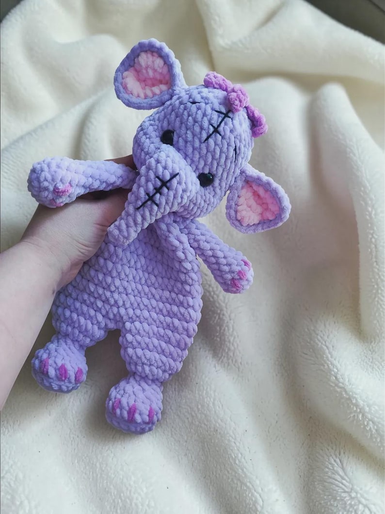 May include: A lavender crochet elephant plush toy with a soft, textured yarn finish. The elephant features large ears with light pink inner details, black safety eyes, a trunk with black stitched accents, and small pink stitched details on its feet.