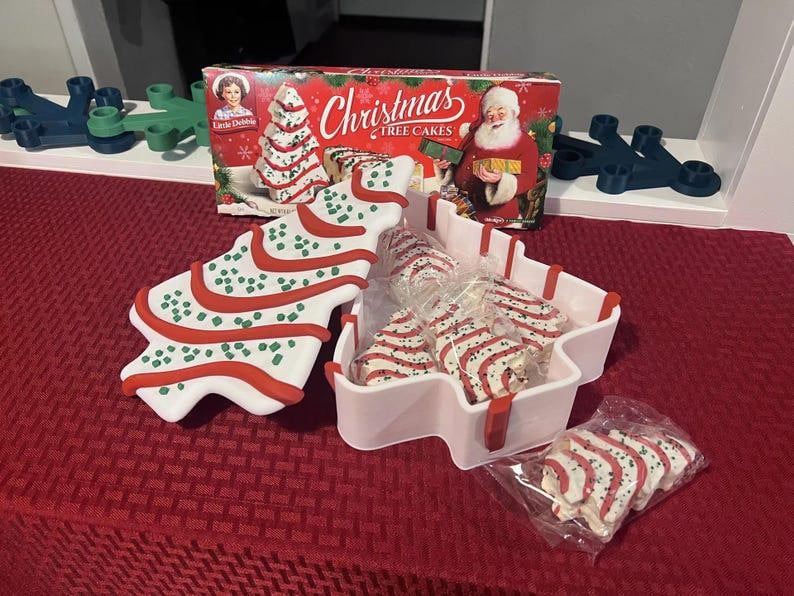 May include: A festive image featuring Christmas Tree Cakes, a white tree-shaped serving dish, and a box of Little Debbie Christmas Tree Cakes. The cakes are white with red stripes and green sprinkles. The box features Santa Claus.