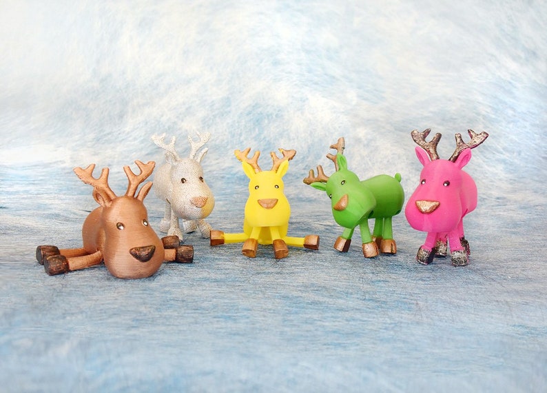 May include: Five colorful reindeer figurines in brown, white, yellow, green, and pink. Each has antlers and a nose in a contrasting color. The reindeer are posed in various positions, some standing and one lying down.