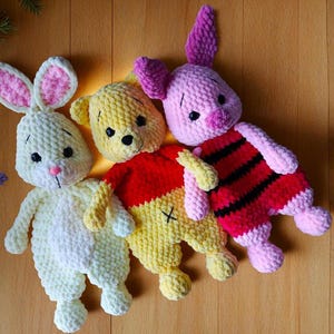 May include: Three plush crochet toys made of soft yarn. On the left, a cream-colored rabbit with pink inner ears and a pink nose. In the center, a yellow bear wearing a red shirt. On the right, a pink piglet with dark horizontal stripes.