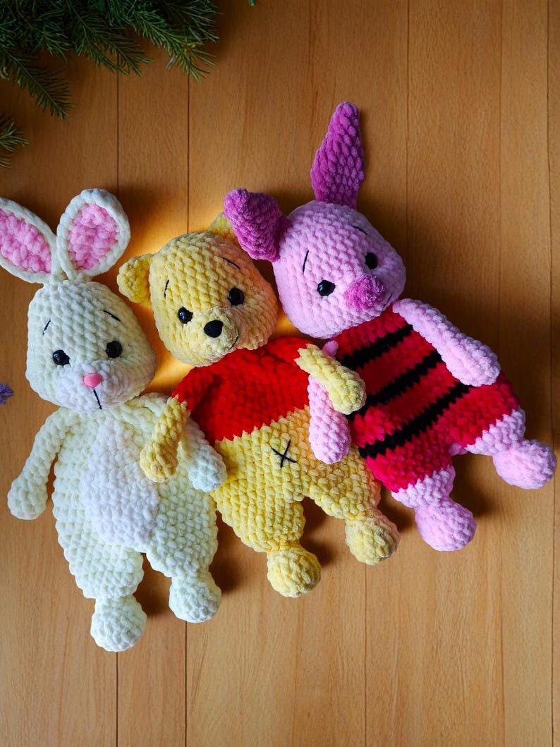 May include: Three plush crochet toys made of soft yarn. On the left, a cream-colored rabbit with pink inner ears and a pink nose. In the center, a yellow bear wearing a red shirt. On the right, a pink piglet with dark horizontal stripes.