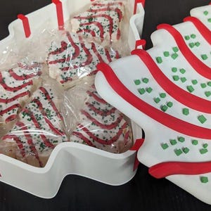 May include: A white, tree-shaped container filled with individually wrapped Christmas tree-shaped cookies. The cookies are decorated with red stripes and green sprinkles. A matching tree-shaped plate is next to the container.