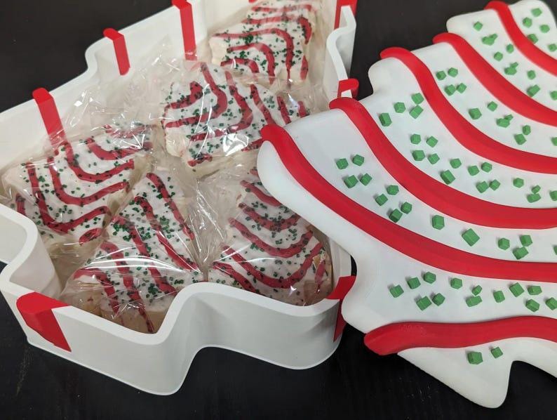 May include: A white, tree-shaped container filled with individually wrapped Christmas tree-shaped cookies. The cookies are decorated with red stripes and green sprinkles. A matching tree-shaped plate is next to the container.