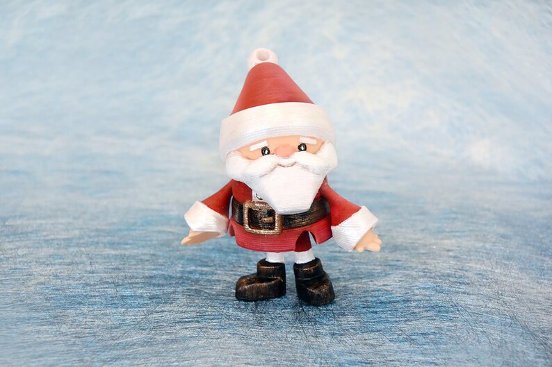 May include: A small Santa Claus figurine with a red suit, white beard, and a matching hat. The figure has a black belt with a gold buckle and black boots. The background is a light blue, textured surface.