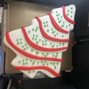 May include: A Christmas tree-shaped cookie with white icing, red icing stripes, and green sprinkles. The cookie is on a printer bed.