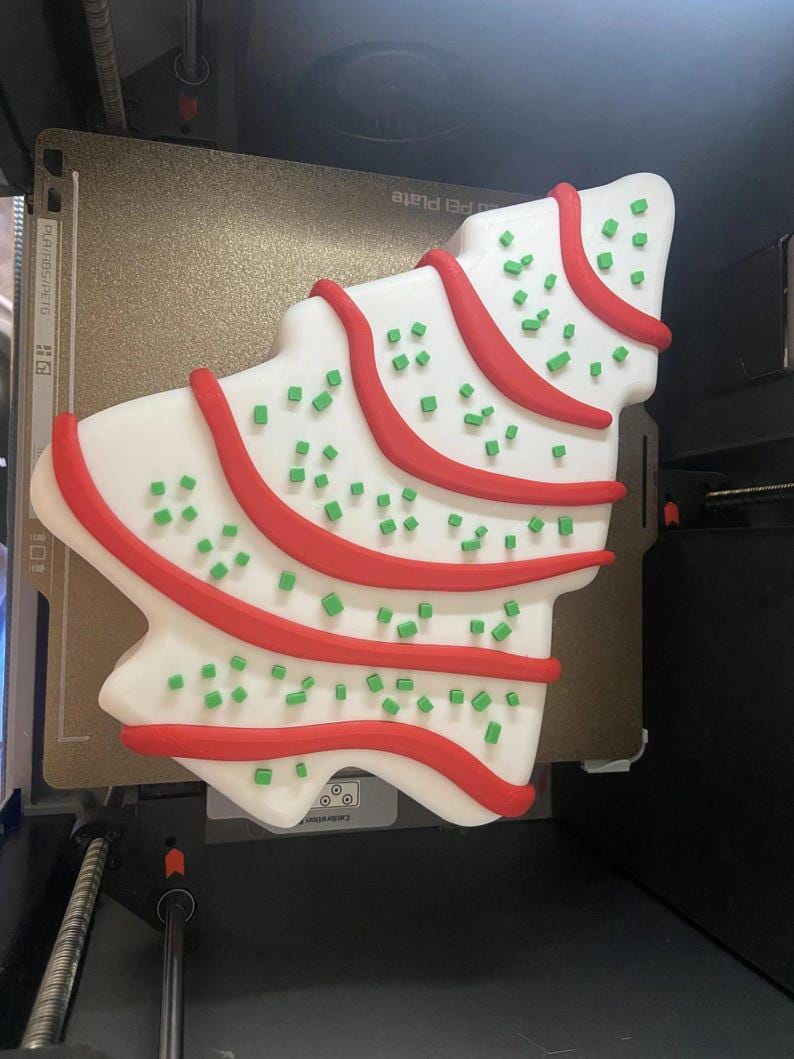 May include: A Christmas tree-shaped cookie with white icing, red icing stripes, and green sprinkles. The cookie is on a printer bed.