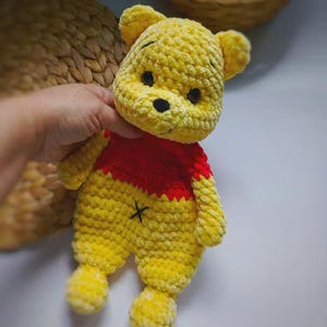 May include: A handmade crochet plush toy featuring a yellow bear character with a red shirt. The bear has black embroidered eyes, a black nose, and a small black 'x' stitch on its yellow belly. The texture is soft and fuzzy with visible yarn loops.
