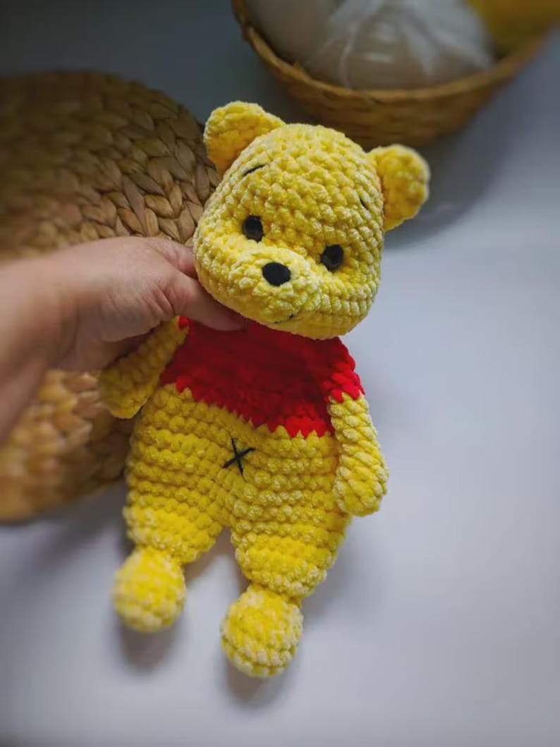 May include: A handmade crochet plush toy featuring a yellow bear character with a red shirt. The bear has black embroidered eyes, a black nose, and a small black 'x' stitch on its yellow belly. The texture is soft and fuzzy with visible yarn loops.