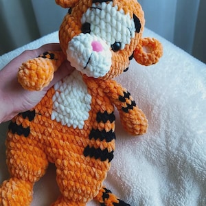 May include: A plush tiger toy made of orange, white, and black yarn. The stuffed animal features a round head with small ears, a white muzzle with a pink nose, and black stripes on its body, limbs, and tail. The texture is soft and fuzzy.