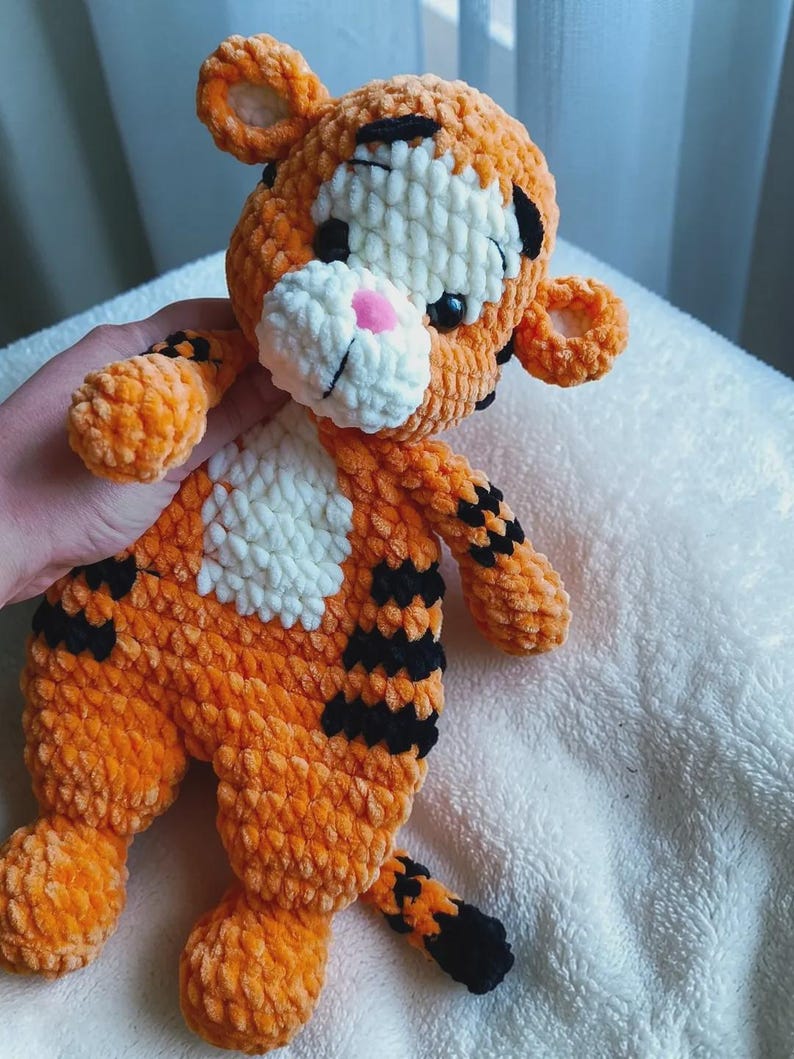 May include: A plush tiger toy made of orange, white, and black yarn. The stuffed animal features a round head with small ears, a white muzzle with a pink nose, and black stripes on its body, limbs, and tail. The texture is soft and fuzzy.