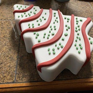 May include: A white, Christmas tree-shaped object with red trim and small green square decorations. The object is sitting on a brown and tan tiled surface.