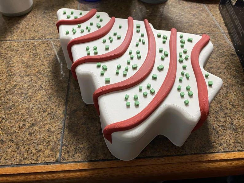 May include: A white, Christmas tree-shaped object with red trim and small green square decorations. The object is sitting on a brown and tan tiled surface.