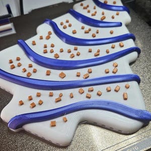 May include: A 3D-printed Christmas tree design in white with blue accents. The tree features blue stripes and small, brown, cube-shaped decorations. The design is on a textured, metallic surface.
