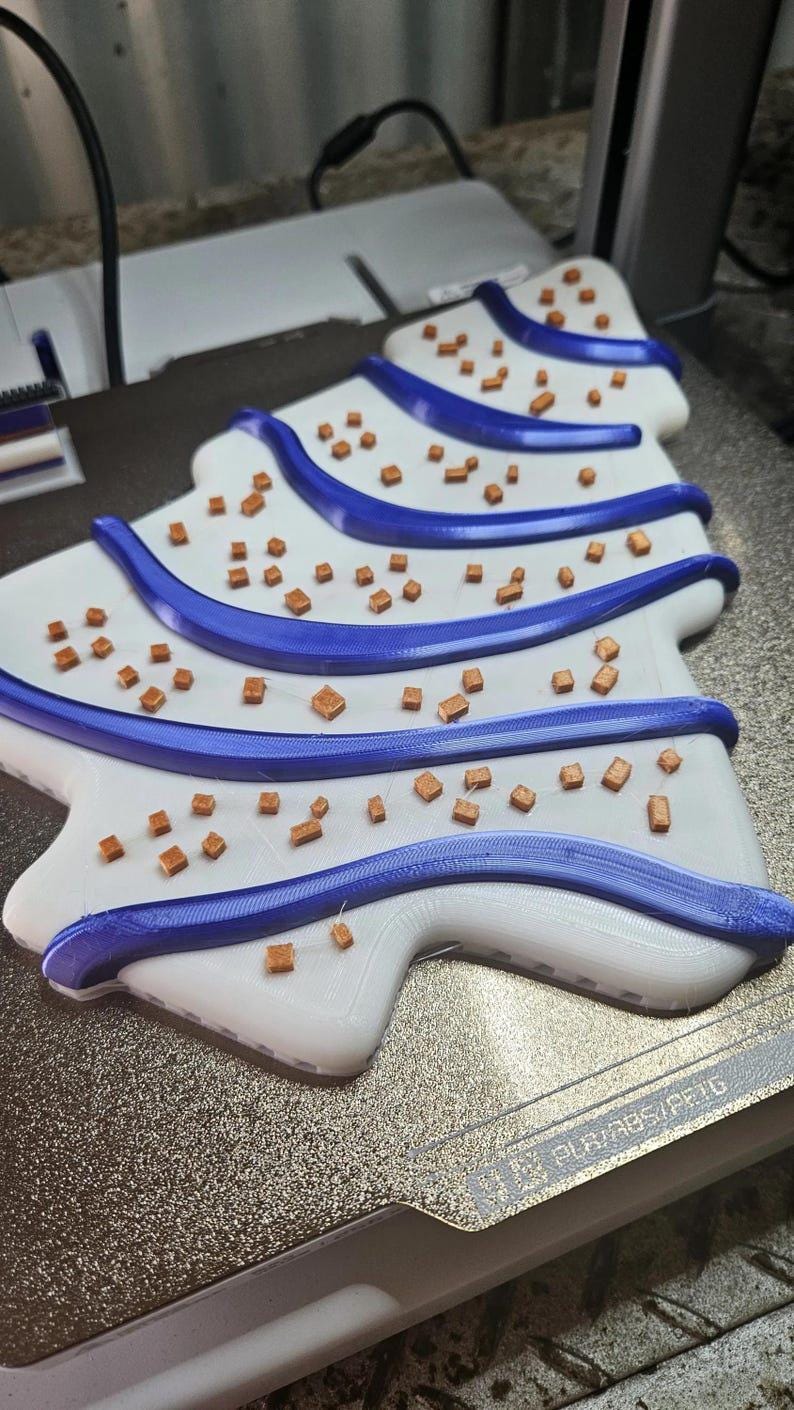 May include: A 3D-printed Christmas tree design in white with blue accents. The tree features blue stripes and small, brown, cube-shaped decorations. The design is on a textured, metallic surface.